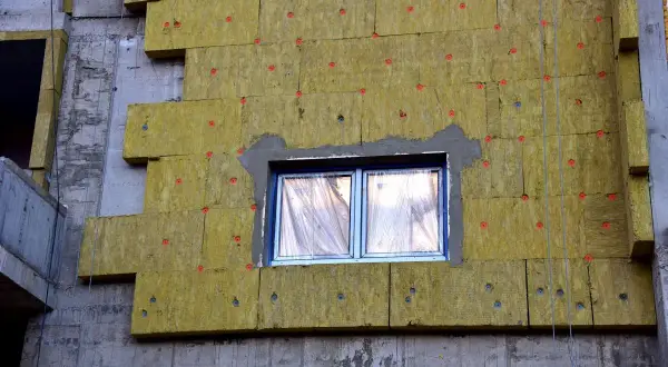 Pannelli isolanti gialli fissati con tasselli rossi su un edificio in costruzione. Si notano alcuni difetti cappotto termico, come fughe e irregolarità.