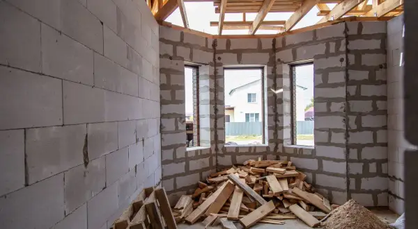 Interno di una casa in costruzione con muri in blocchi di cemento, travi in legno a vista e cumuli di legname. Sembra un cantiere fermo da tempo, quasi un abuso edilizio dopo 20 anni.