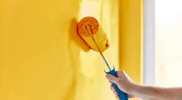 Mano che imbianca un muro giallo con un rullo. La temperatura ideale per imbiancare è tra i 10°C e i 25°C per una buona asciugatura.
