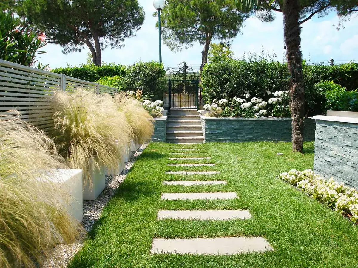 Un bellissimo giardino progetto con un sentiero di lastre di pietra, erba verde, cespugli fioriti e un muretto in pietra.