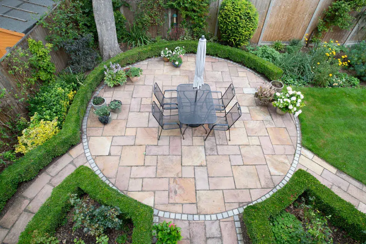 Un'elegante pavimentazione esterna in pietra circonda un tavolo da pranzo con ombrellone, immerso in un giardino curato con siepi geometriche e aiuole fiorite.