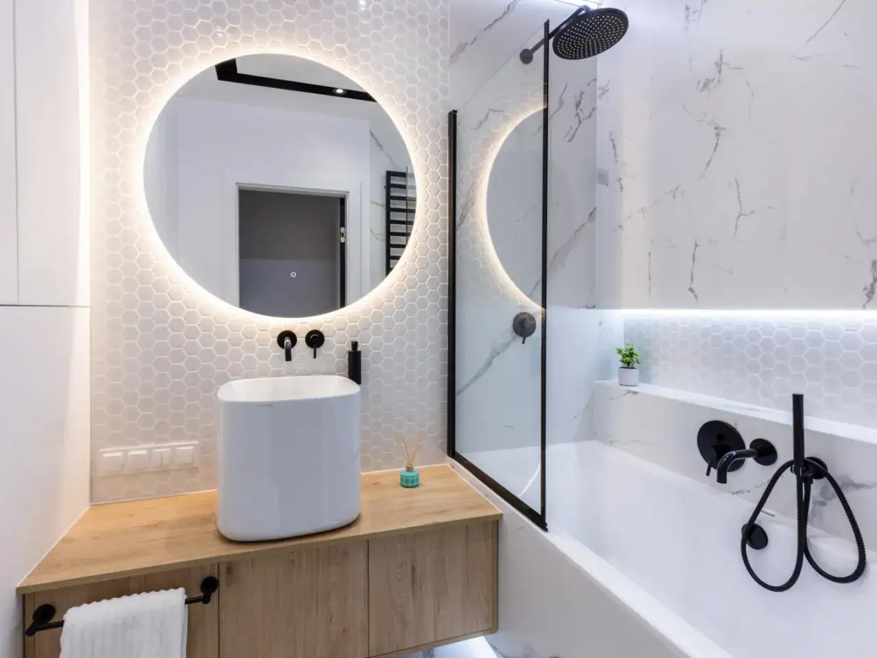 Elegante bagno senza finestra con lavabo bianco su mobile in legno, specchio rotondo illuminato e vasca con doccia.