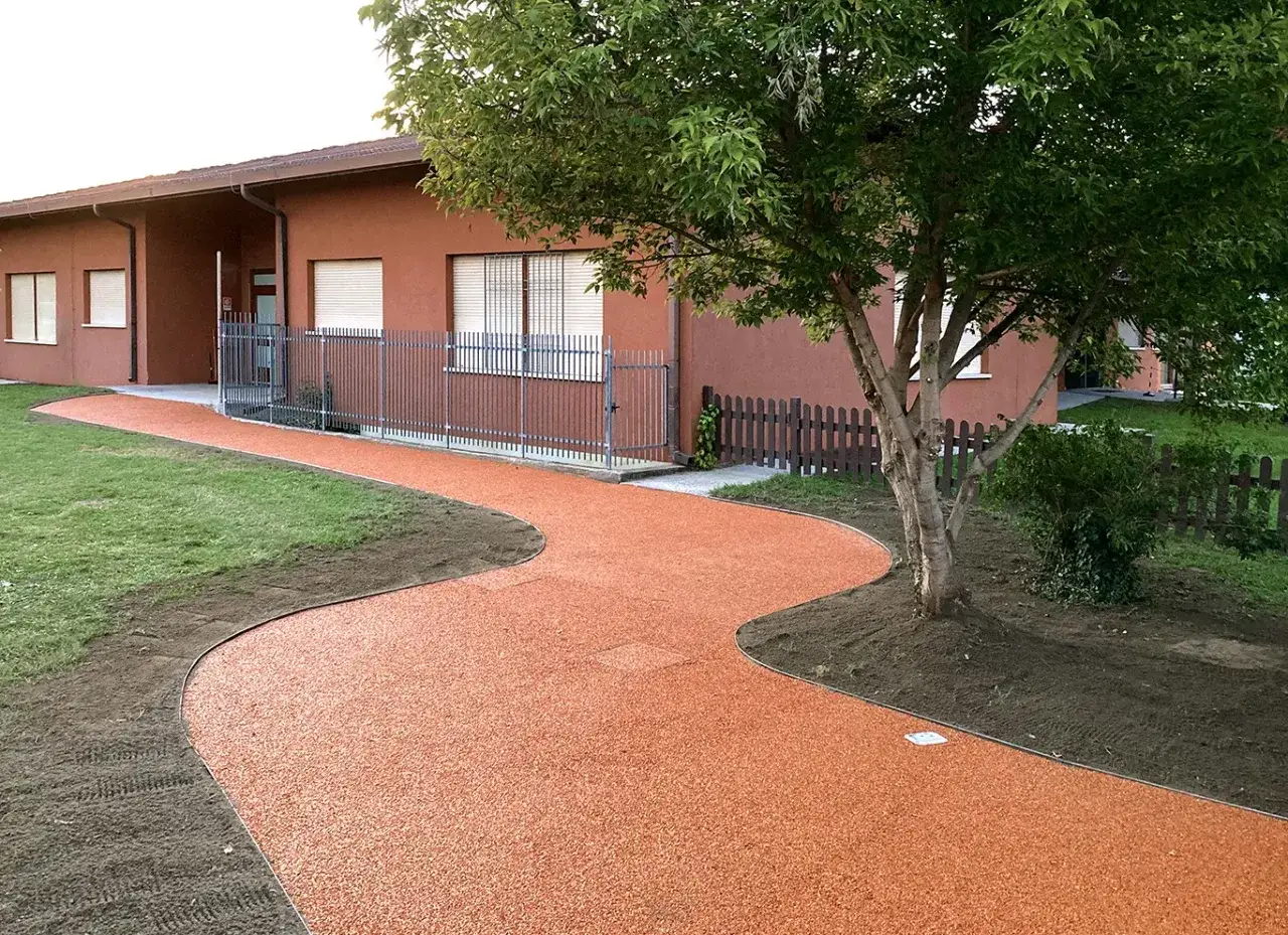 Un sinuoso vialetto carrabile in giardino, con ghiaia arancione, conduce a un edificio con facciata color terra e finestre con persiane.