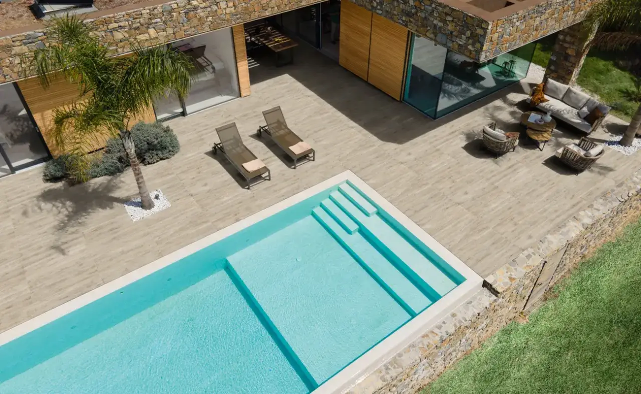 Piscina e area relax su una moderna pavimentazione esterna in legno. Due lettini prendisole e mobili da giardino completano lo spazio.