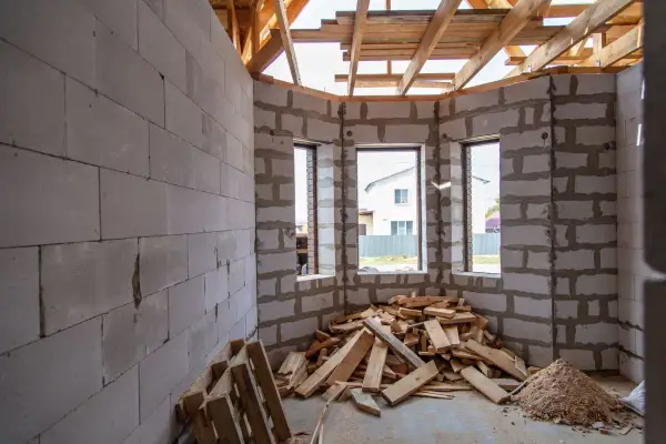 Interno di una casa in costruzione con muri in blocchi di cemento, travi in legno a vista e cumuli di legname. Sembra un cantiere fermo da tempo, quasi un abuso edilizio dopo 20 anni.